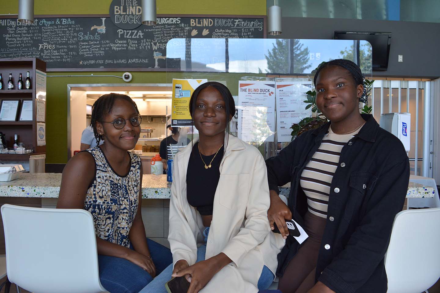 Three students sitting down, in the cafeteria, smiling at the camera